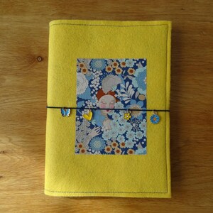 Op de afbeelding: Een gele vilten tijdschriftomslag met een bloemenpatroon en een zwarte elastische band. De band is versierd met vlinder- en bloemhangers. De tijdschriftomslag ligt op een houten oppervlak.