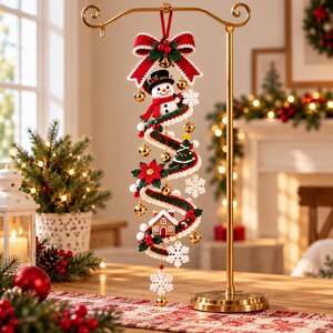 May include: A vertical crochet Christmas ornament hanging from a gold stand. It features a red bow, a snowman, a small Christmas tree, a gingerbread house, white snowflakes, green and red garland, and small gold jingle bells.