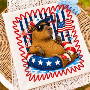 May include: A white fabric square featuring a brown capybara wearing a blue and white baseball cap and black sunglasses. The capybara sits in a red, white, and blue star-patterned inner tube, holding a small American flag against a blue striped background.