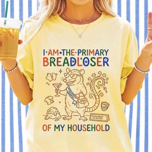 May include: A pale yellow t-shirt featuring a graphic of an opossum wearing sunglasses and a sash that reads BREADLOSER CHAMPION. The text I AM THE PRIMARY BREADLOSER OF MY HOUSEHOLD is printed in colourful letters alongside illustrations of a martini glass, a purse, and a broken piggy bank.