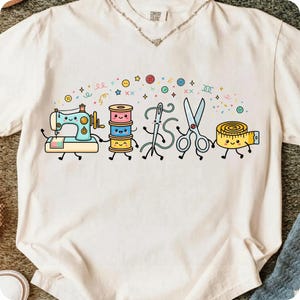 May include: An off-white t-shirt featuring a graphic print of anthropomorphic sewing supplies including a light blue sewing machine, a stack of colorful thread spools, a needle, scissors, and a yellow measuring tape, all with smiling faces.