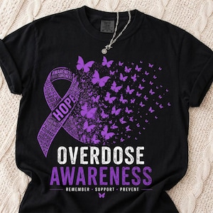Op de afbeelding: Een zwart t-shirt met een paars bewustzijnslint bestaande uit woorden als hoop, kracht en moed. Het lint gaat over in een zwerm paarse vlinders. Daaronder staat de tekst OVERDOSE AWARENESS REMEMBER SUPPORT PREVENT.