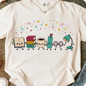 May include: A cream-colored short-sleeve t-shirt featuring a graphic of anthropomorphic reading-themed items including an open book, a stack of three books, a coffee mug, a bookmark, eyeglasses, and a desk lamp, all surrounded by colorful confetti.