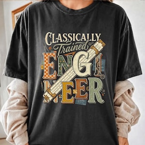 Może przedstawiać: Ciemnoszary t-shirt z napisem "CLASSICALLY Trained ENGINEER" w czcionce w stylu vintage. Grafika zawiera ukośną linijkę nałożoną na litery oraz małe ilustracje przyborów kreślarskich i kół zębatych.