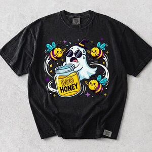 May include: A black short-sleeve t-shirt featuring a graphic of a white ghost wearing sunglasses and a witch hat, holding a jar of honey labeled BOO HONEY. The ghost is surrounded by three angry-looking yellow and black striped bees and stars.