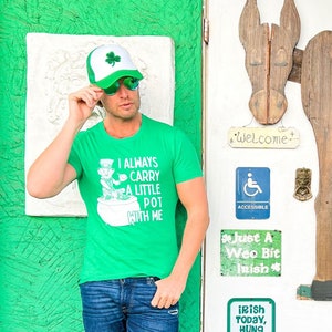 May include: A man wearing a green t-shirt with a shamrock and the text "I always carry a little pot with me" and a green and white baseball cap. He is standing in front of a white door with a wooden donkey cutout and signs that say "Welcome", "Just a wee bit Irish", and "Irish today, hung over tomorrow!" The door is on a green wall with a small white statue of a donkey on the ground.