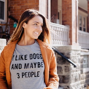 May include: A woman wearing a gray t-shirt with the text "I LIKE DOGS AND MAYBE 3 PEOPLE" and a rust-colored coat. She is standing on a stone staircase in front of a brick building.