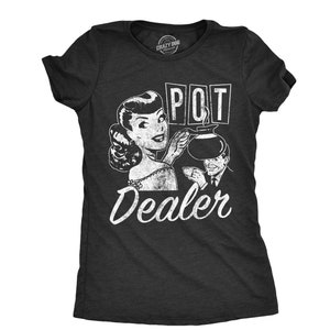 Puede incluir: Una camiseta gris oscuro con una ilustración de estilo vintage de una mujer vertiendo café de una olla. El texto "POT DEALER" está impreso en blanco en la camiseta.