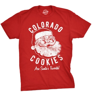 May include: Red t-shirt with a white distressed graphic of Santa Claus and the text "Colorado Cookies Are Santa's Favorite!"