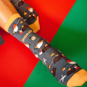 Könnte beinhalten: Ein Paar blaue Socken mit gelbem Bündchen und einem Gnom-Muster. Die Socken zeigen einen Gnom, der einen Koffer trägt, und das Wort "GNOMAD" ist auf der Socke aufgedruckt. Die Socken befinden sich auf einem roten und grünen Hintergrund.