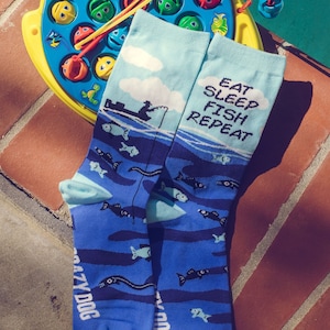 May include: A pair of blue socks with a fishing theme. The socks feature a cartoon fisherman in a boat, fish, and the text "Eat Sleep Fish Repeat".