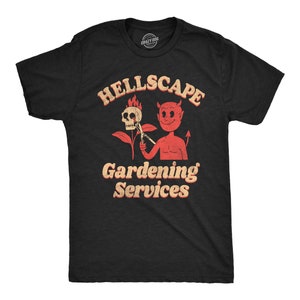 Puede incluir: Camiseta negra con un diablo rojo sosteniendo una calavera y una herramienta de jardinería. El texto "Hellscape Gardening Services" está impreso en naranja en la camiseta.