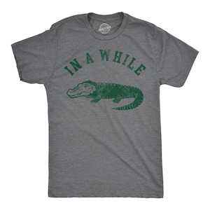 May include: A heather grey t-shirt with a green alligator graphic and the text "In a While" printed on the front.