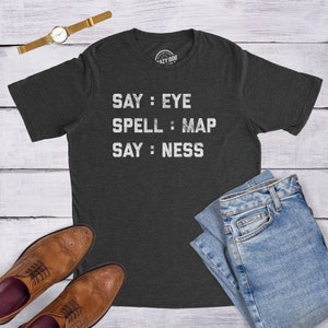 May include: A dark heather grey t-shirt with the text "SAY: EYE SPELL: MAP SAY: NESS" printed in white.