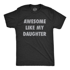 May include: A black t-shirt with the text "AWESOME LIKE MY DAUGHTER" in white, distressed lettering. The shirt has short sleeves and a crew neck. The Crazy Dog brand logo is visible at the top.