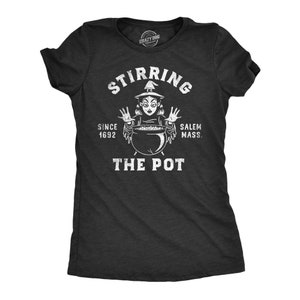 Pode incluir: Uma camiseta preta com um gráfico branco de uma bruxa mexendo um caldeirão. O texto diz "Stirring the Pot" com "Since 1692 Salem Mass." abaixo.
