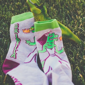 Puede incluir: Un par de calcetines blancos con detalles en verde y rosa. Un calcetín tiene la palabra "Bridezilla" en letras verdes. El otro calcetín tiene un dinosaurio verde con un vestido de novia blanco.
