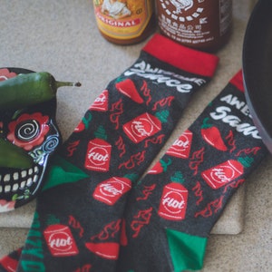 Könnte beinhalten: Ein Paar schwarze Socken mit einem roten, grünen und weißen Design. Die Socken haben ein sich wiederholendes Muster aus scharfen Saucenflaschen mit dem Wort "Hot" darauf. Die Socken haben auch den Text "Awesome Sauce" und "Spicy Dog" darauf.