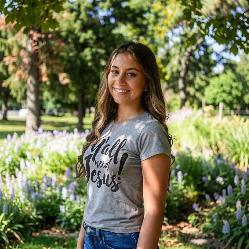 May include: A gray t-shirt with the text "Y'all need Jesus!" in black script. The shirt is short-sleeved and has a crew neck. The person is wearing blue jeans. The background is a park with trees and flowers.
