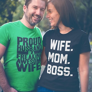 May include: A bright green t-shirt with black text reading PROUD HUSBAND OF A FREAKING AWESOME WIFE and a black t-shirt with white text reading WIFE. MOM. BOSS. Both shirts feature short sleeves and a crew neckline, made of a cotton-like fabric.