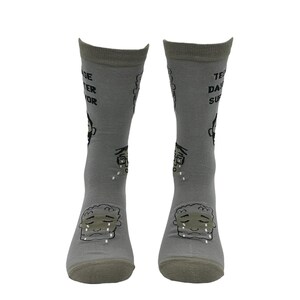 May include: A pair of gray socks with a cartoon illustration of a crying face on the front. The socks have text that reads "Teenage Disaster Survivor" and other graphics. The socks are made of a soft material.