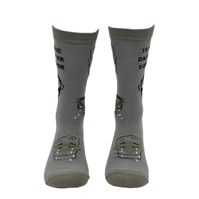 May include: A pair of gray socks with a cartoon illustration of a crying face on the front. The socks have text that reads "Teenage Disaster Survivor" and other graphics. The socks are made of a soft material.