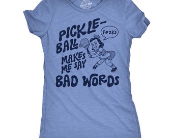 May include: Light blue t-shirt with the text "PICKLEBALL MAKES ME SAY BAD WORDS" in navy blue. A cartoon illustration of a girl playing pickleball is also featured. The shirt has a crew neck and short sleeves.