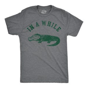Op de afbeelding: Een antracietgrijs T-shirt met korte mouwen en een donkergroene afbeelding van een alligator in het midden op de borst. Boven de alligator staat de tekst "IN A WHILE" in een verweerd, donkergroen, gebogen lettertype. Het shirt heeft een ronde hals.