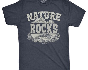 May include: A dark navy blue short-sleeve t-shirt featuring a graphic print in light gray. The design displays the text "NATURE ROCKS" in a bold, distressed font above an illustration of a rocky shoreline with clouds in the sky.