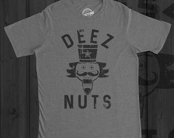May include: A heather grey short-sleeve t-shirt featuring a black distressed graphic of a nutcracker wearing a top hat with a star. The text DEEZ NUTS is printed in a distressed black font above and below the nutcracker illustration.