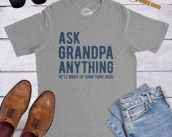May include: A heather gray short-sleeve t-shirt with dark blue text that reads ASK GRANDPA ANYTHING HE'LL MAKE UP SOMETHING GOOD. The shirt is displayed with black sunglasses, a gold-tone wristwatch, and blue denim jeans.