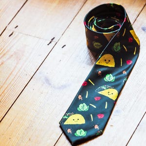 May include: A black necktie featuring a repeating pattern of yellow taco illustrations with smiling faces, interspersed with small green lettuce leaves, red tomato pieces, and thin yellow strips of shredded cheese scattered across the fabric.