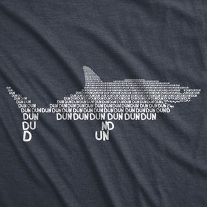 Puede incluir: Una camiseta azul oscuro con una silueta de tibur&oacute;n blanca hecha con la palabra "dun" repetida muchas veces. Las palabras "du", "d", "nd" y "un" tambi&eacute;n son visibles en la camiseta.