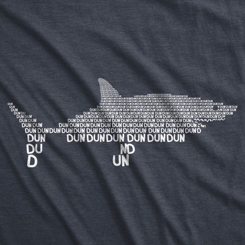 Puede incluir: Una camiseta azul oscuro con una silueta de tibur&oacute;n blanca hecha con la palabra "dun" repetida muchas veces. Las palabras "du", "d", "nd" y "un" tambi&eacute;n son visibles en la camiseta.