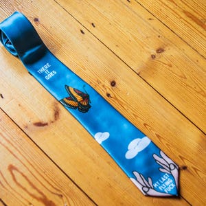 May include: A blue tie featuring a butterfly and cloud pattern. The tie displays the text "THERE IT GOES" and "MY LAST FLYING FUCK". It rests on a wooden surface.