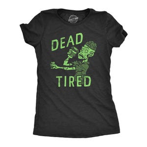 May include: A charcoal gray short-sleeve t-shirt featuring a light green graphic of a zombie holding a coffee cup. The text "DEAD" appears above the zombie and "TIRED" appears below in a distressed, light green, sans-serif font style.