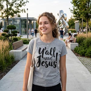 May include: A gray t-shirt with the text "Y'all need Jesus" in black script. The shirt is worn by a person walking on a paved path in a park setting. The person is smiling and carrying a beige tote bag.