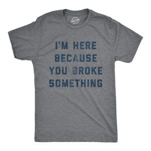 May include: A heather gray short-sleeve t-shirt with a crew neckline. The front features dark blue, distressed-style text that reads: I'M HERE BECAUSE YOU BROKE SOMETHING. The fabric has a visible heathered texture throughout the entire garment.