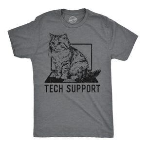 May include: A dark heather gray short-sleeve t-shirt featuring a black graphic of a fluffy cat sitting on a laptop. Below the laptop, the text TECH SUPPORT is printed in a bold, distressed black font. The shirt has a crew neckline.