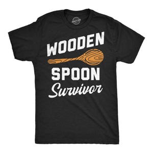 Puede incluir: Camiseta negra con las palabras "WOODEN SPOON Survivor" en blanco y un gráfico de cuchara de madera naranja. La camiseta tiene cuello redondo y mangas cortas. El logo de la marca "Crazy Dog" es visible en la parte superior de la espalda.