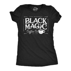 Può includere: Una t-shirt nera a maniche corte con testo bianco dall'aspetto usurato che recita "BLACK MAGIC Coffee Co", con "SALEM, MASSACHUSETTS" sopra e "EST. 1690" sotto. Un piccolo grafico di una tazza di caffè fumante è posto accanto alla parola Coffee.