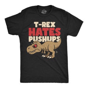 Op de afbeelding: Een zwart t-shirt met korte mouwen en een afbeelding van een beige T-Rex met rode ogen. Boven de dinosaurus staat de tekst T-REX HATES PUSHUPS in dikke rode en crèmekleurige letters. Het ontwerp is gecentreerd op de borst van het kledingstuk.