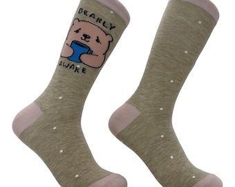 May include: A pair of heathered tan crew socks with light pink cuffs, heels, and toes. The socks feature small white polka dots throughout. One sock displays a graphic of a light brown bear holding a blue mug with the text BEARLY AWAKE.