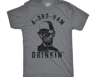 May include: A charcoal grey heather t-shirt featuring a black graphic of Abraham Lincoln wearing a baseball cap and sunglasses with an American flag pattern. The text A-BRO-HAM DRINKIN' is printed in black above and below the portrait.