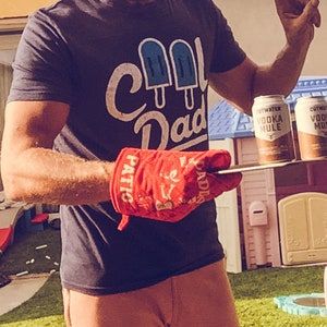 Op de afbeelding: Een persoon draagt een blauw T-shirt met de tekst "Cool Dad" en een afbeelding van drie blauwe ijslolly's. De persoon houdt een rode ovenwant vast met de tekst "Patriotic" en "Paddles" erop. De persoon staat voor twee blikjes Cutwater Vodka Mule.