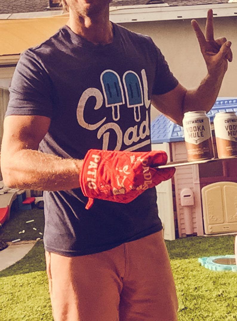 Op de afbeelding: Een persoon draagt een blauw T-shirt met de tekst "Cool Dad" en een afbeelding van drie blauwe ijslolly's. De persoon houdt een rode ovenwant vast met de tekst "Patriotic" en "Paddles" erop. De persoon staat voor twee blikjes Cutwater Vodka Mule.