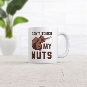 Könnte beinhalten: Weiße Keramiktasse mit einem braunen Eichhörnchen, das eine Waffe hält, und dem Text "Don't Touch My Nuts" in braunen Buchstaben.