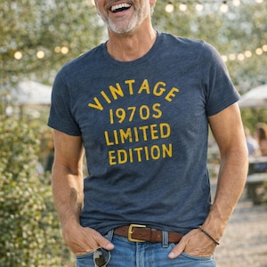 May include: A heathered navy blue short-sleeve t-shirt featuring the yellow text VINTAGE 1970S LIMITED EDITION printed on the chest. The shirt is paired with blue denim jeans, a brown leather belt with a gold buckle, and a beaded bracelet.