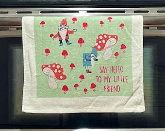 Peut inclure: Un torchon de cuisine vert clair avec un motif répétitif de champignons rouges et blancs et deux gnomes. Le texte imprimé en rouge indique, Say hello to my little friend. Le torchon a une bordure blanche et est posé sur un four noir.