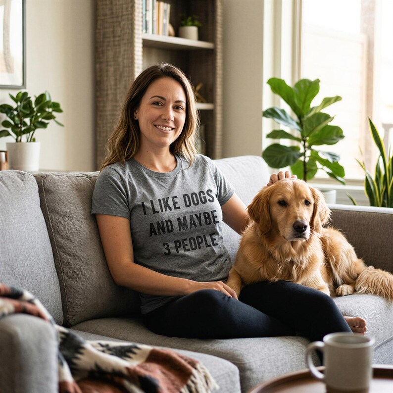 May include: A woman and a golden retriever dog are sitting on a gray sofa. The woman is wearing a gray t-shirt with the text "I LIKE DOGS AND MAYBE 3 PEOPLE". A mug sits on a nearby table.